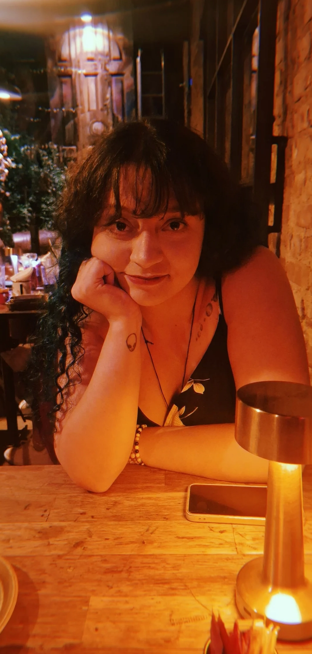 A fotografia mostra uma mulher jovem sentada a uma mesa de madeira em um ambiente com iluminação quente e aconchegante, que parece ser um restaurante ou bar.Ela está em primeiro plano, olhando diretamente para a câmera com uma expressão serena e um leve sorriso.A mulher tem pele clara, cabelos escuros, longos e ondulados, com uma franja sobre a testa. Algumas mechas de seu cabelo parecem ter uma tonalidade azulada ou esverdeada. Seus olhos são escuros e expressivos.Ela apoia o rosto na mão direita, com o cotovelo sobre a mesa. Nesta mão, há uma pequena tatuagem de um contorno que lembra a cabeça de um alienígena. Ela veste uma blusa escura de alças com uma estampa sutil.À sua frente, sobre a mesa, há um pequeno abajur moderno e sem fio, de cor metálica, que ilumina seu rosto e o ambiente ao redor com uma luz amarelada. Ao lado do abajur, há um prato, talheres e um celular.O fundo está ligeiramente desfocado. À direita, uma parede de tijolos aparentes compõe o cenário. À esquerda, outras pessoas estão sentadas em mesas mais distantes, sugerindo um local movimentado, mas a atmosfera geral da foto é íntima e focada na mulher.